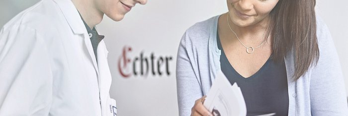 Das Bild zeigt einen Mann mit weißem Kittel, der zusammen mit einer Frau auf ein Klemmbrett in seinem Händen schaut