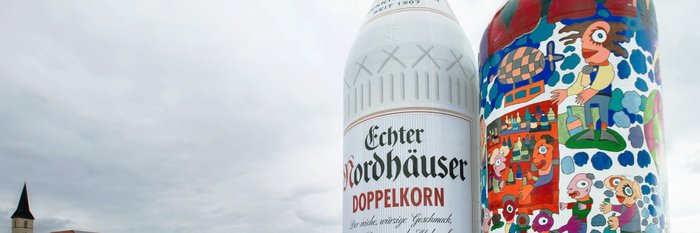 Das Bild zeigt zwei riesige Flaschen: eine Nordbrand Nordhausen Doppelkorn Flasche und eine Bunt gestaltete Flasche auf dem Werksgelände von Nordbrand