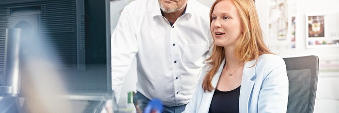Das Foto zeigt eine junge Frau im Büro, neben ihr steht ein Mann. Beide schauen auf einen Bildschirm