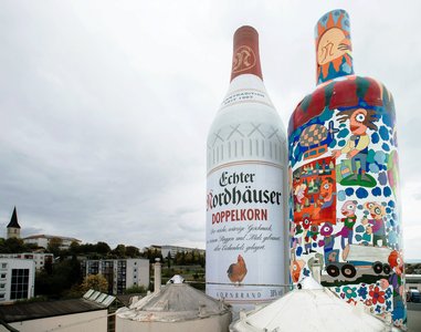 Das Bild zeigt zwei riesige Flaschen: eine Nordbrand Nordhausen Doppelkorn Flasche und eine Bunt gestaltete Flasche auf dem Werksgelände von Nordbrand