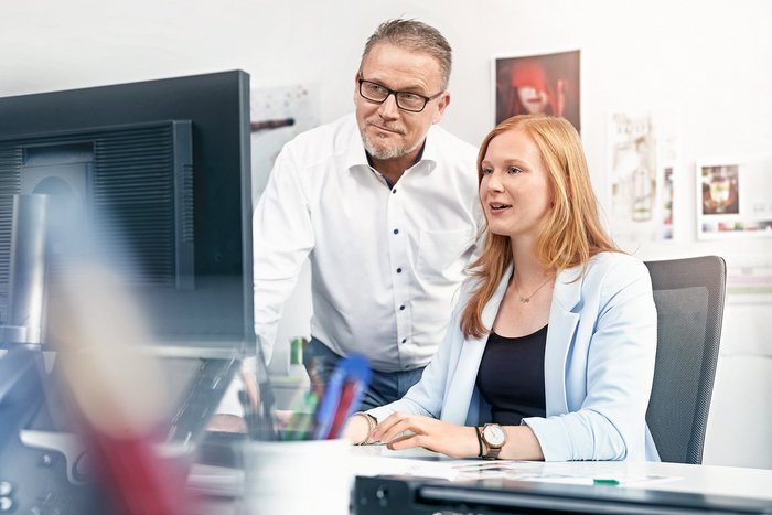 Das Foto zeigt eine junge Frau im Büro, neben ihr steht ein Mann. Beide schauen auf einen Bildschirm